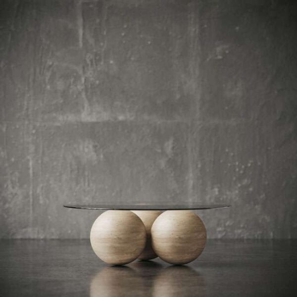 Modern round coffee table featuring a glass top supported by three light wooden spheres, placed on a concrete floor against a grey wall. طاولة قهوة دائرية عصرية تحتوي على سطح زجاجي مدعوم بثلاث كرات خشبية فاتحة اللون، موضوعة على أرضية خرسانية أمام جدار رمادي.