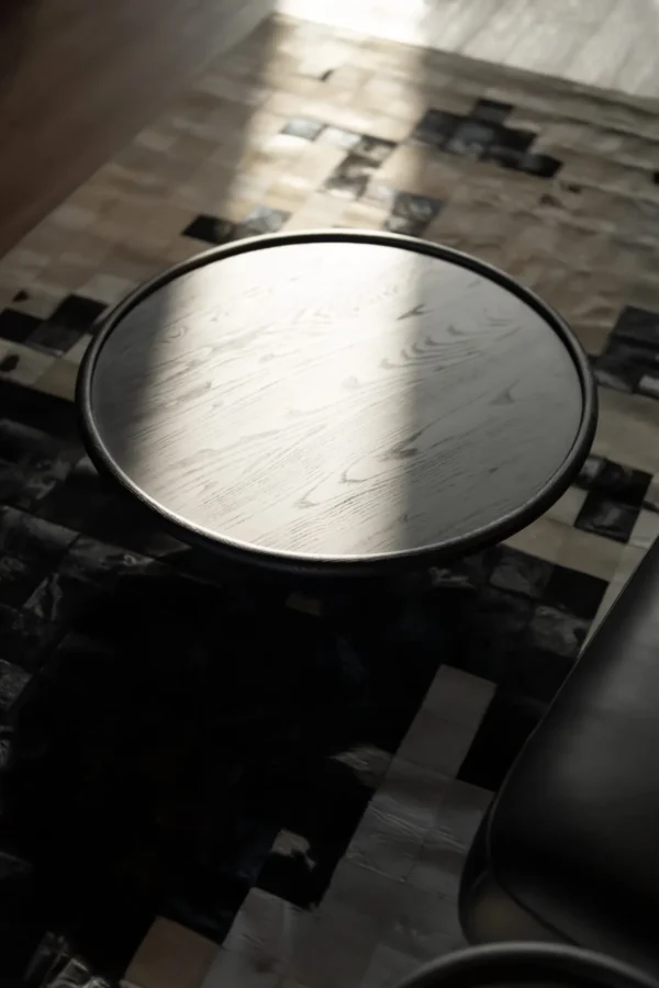 The Nocturne Coffee Table with a round black wooden top illuminated by soft natural light, resting on a patterned neutral rug. The photo highlights the table’s elegant grain texture and minimal silhouette within a warm, modern living space.