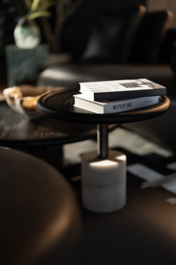 A close-up of the smaller Nocturne table featuring a dark wooden circular top and a white marble base, styled with minimalist books. Warm natural light enhances the table’s elegant materials and soft modern design.