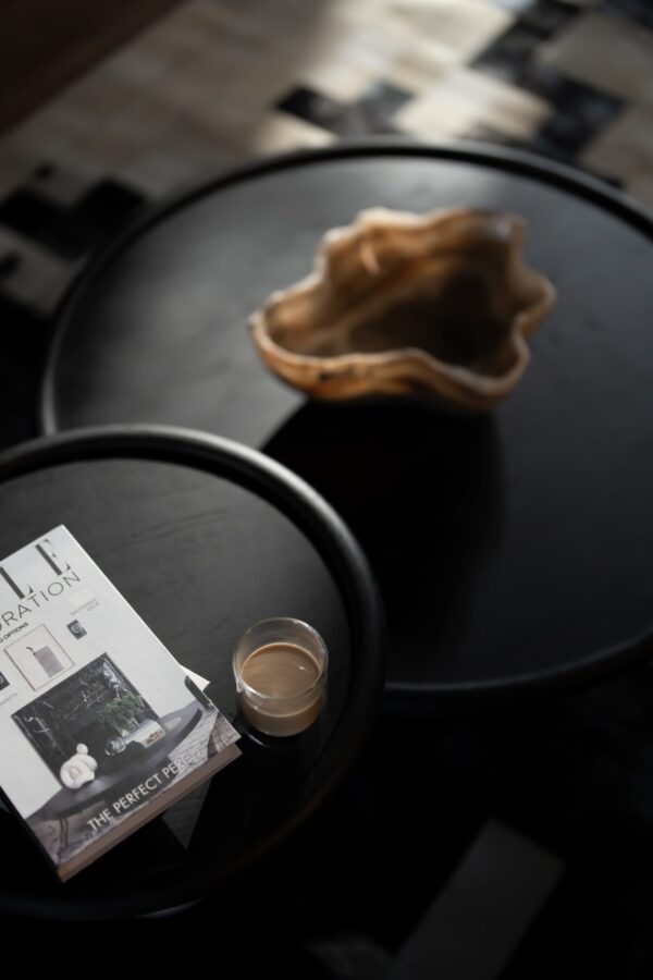 A styled view of the Nocturne Coffee Table set, featuring a dark wooden surface with a sculptural bowl, stacked magazines, and a glass of coffee. Soft daylight highlights the table’s smooth texture and refined craftsmanship.