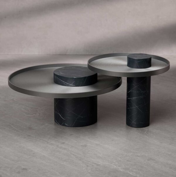 A composition of two Bravura Gemina Coffee Tables, featuring contrasting heights and a circular design. The tables combine dark marble cylindrical bases with brushed metal tops, creating a bold architectural balance.