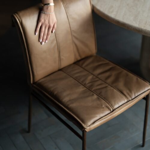 Bon Appétit Dining Chair — a modern tan leather dining chair with a softly cushioned seat and backrest, paired with slim bronze legs. Captured beside a natural stone table, this image highlights tactile luxury and refined craftsmanship.