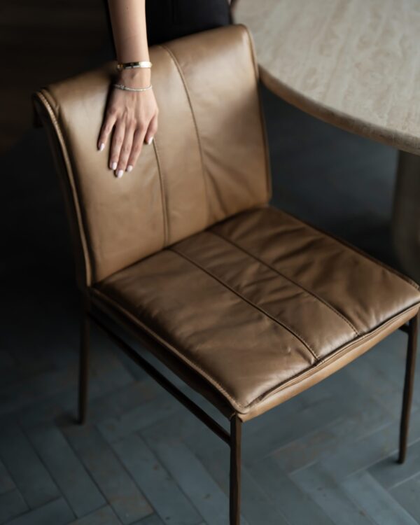 Bon Appétit Dining Chair — a modern tan leather dining chair with a softly cushioned seat and backrest, paired with slim bronze legs. Captured beside a natural stone table, this image highlights tactile luxury and refined craftsmanship.